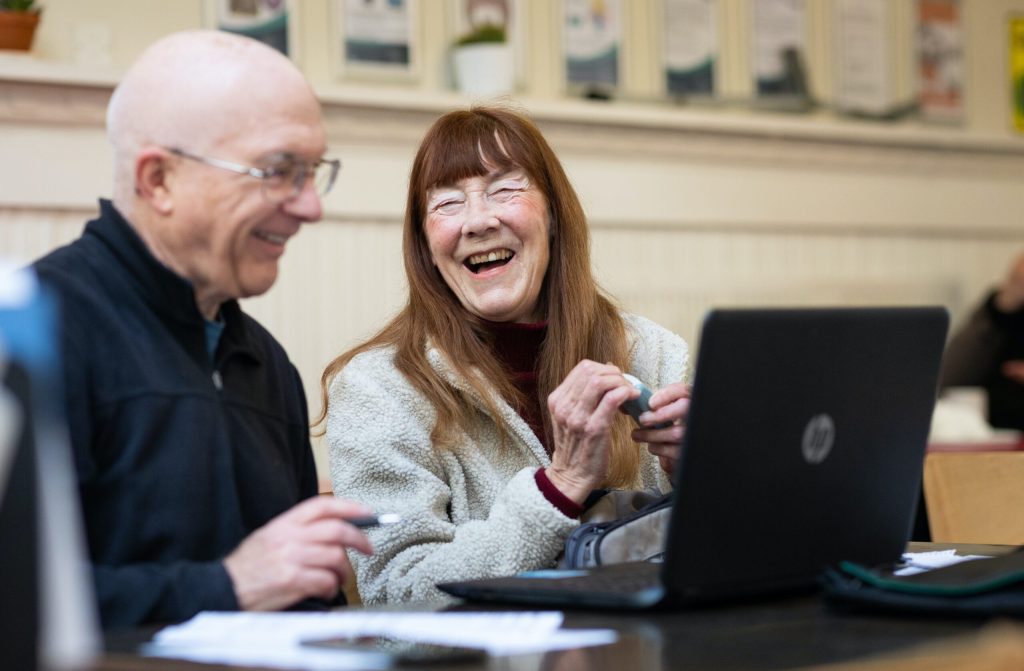 Two older people talk and use a laptop