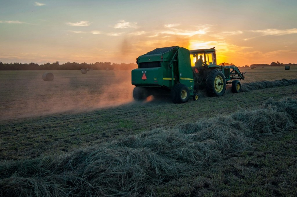 A tractor pulls a hay baler across a field at sunset, kicking up dust as round hay bales rest in the background.
